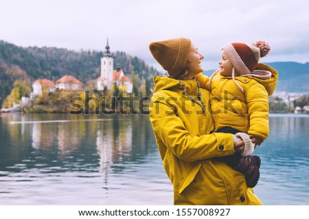 Family Travel Europe. Mother and child in yellow raincoats looking at Bled Lake. Autumn or Winter in Slovenia. View on Island with Church on Bled Lake with Castle and Mountains in Nature Background.