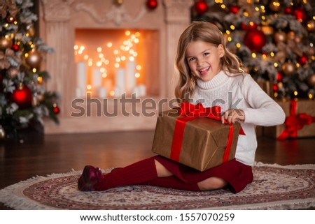 Cute girl waiting for the New Year, sitting at home in front of the fireplace. she is holding a gift with red ribbons, in the background is a Christmas tree with bright lights.