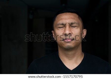 A face of an Asian Indonesian man forcing his eyes closed as if he 's ridden with guilty conscience. The portrait is on black and the model wears black t-shirt Royalty-Free Stock Photo #1557170795