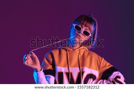 Daring pretty young 20s fashion teen girl model wear glasses chewing bubble gum look at camera stand at purple studio background, igen teenager with bubblegum in trendy night 80s party light portrait Royalty-Free Stock Photo #1557182312
