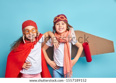 Two joyful caucasian children in pilot suit, cloak and cardboard airplane stand together isolated over blue background
