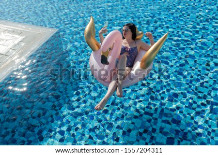 Young woman taking a selfie with mint smartphone on a pink and gold swan / flamingo pool float in the swimming pool on vacation