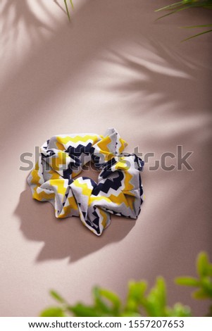 Object photo of a scrunchie with abstract ornament. The scrunchie is lying on a beige background. There are leaves in corners of a photo. The scrunchie and leaves casting a shadow.