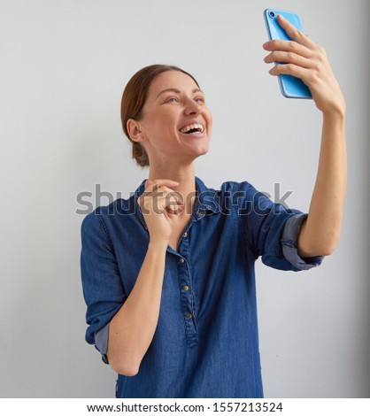 Happy young woman in blue casual jeans shirt dress active listening and laughs with teeth smile and gestures to selfie camera in video chat conference call on phone in hand holds up on white home wall Royalty-Free Stock Photo #1557213524