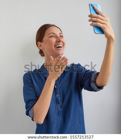 Happy young woman in blue casual jeans shirt dress speak and laughs with teeth smile and gestures to selfie camera in video talk chat conference call on phone in hand holds up on white home wall Royalty-Free Stock Photo #1557213527