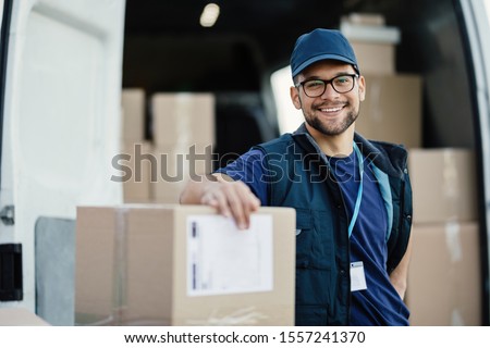 Portrait of happy worker unloading boxes from a delivery van and looking at camera.  Royalty-Free Stock Photo #1557241370