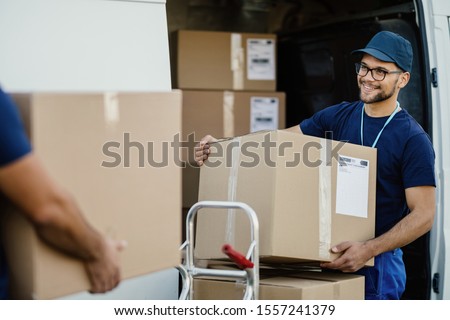 Young happy delivery man unloading boxes from a mini van and talking with his coworker. 