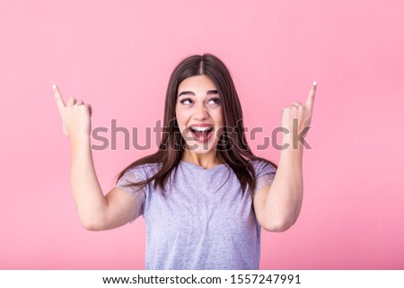 Portrait of brunette woman with long hair in basic t-shirt rejoicing and pointing finger at copyspace isolated over pink background