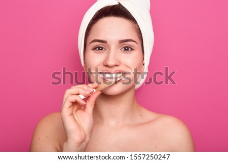 Image of beautiful young girl wrapped white towel, cleaning her teeth, looks happy, being in good mood, doing morning procedures in bathroom, posing with bared shoulders against pink studio wall.