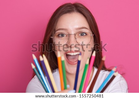 Close up portrait of young caucasian girl with staright hair holding colored pencils while looking directly at camera and keepsmouth opened, model posing isolated over pink studio background.