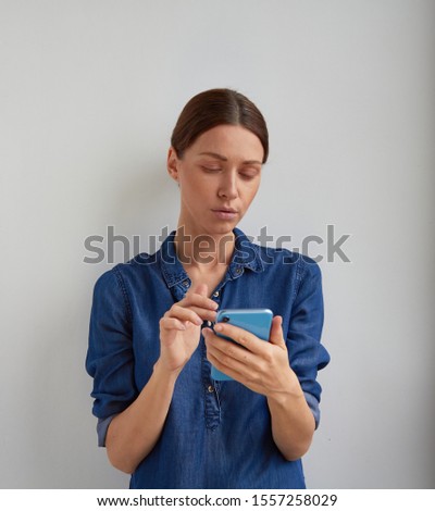 Isolated dissatisfied brunette woman in casual blue jeans shirt dress reads message or looks news feed with sad face holding phone by two hands on white wall studio background Royalty-Free Stock Photo #1557258029