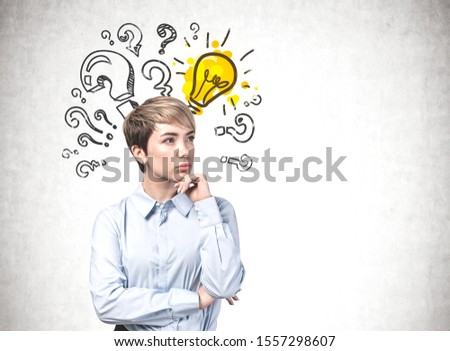 Thoughtful young blonde businesswoman standing near concrete wall with bright yellow lightbulb and question marks drawn on it. Concept of looking for answer. Mock up