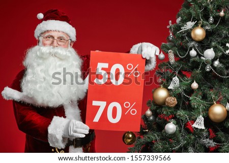 Portrait of senior man in Santa costume holding placard with big sale while standing near the Christmas tree over red background