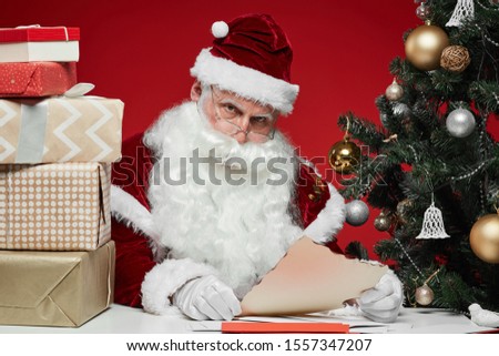 Santa Claus sitting at the table with stack of gift boxes and reading letters from children with Christmas tree in the background