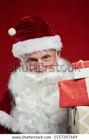 Portrait of Santa Claus in hat holding gift boxes and looking at camera isolated on red background