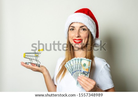 A young woman in a Santa Claus hat holds in her hands a shopping trolley and money on a white background. Christmas sale concept, black friday, discount
