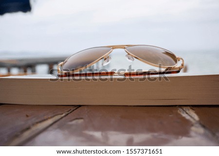 Sunglasses and a book on the table on beach towards sea and coast pier. Holiday, vacation and relaxation conceptual photography. Low view of a retro or modern sunglasses at seaside or shore.