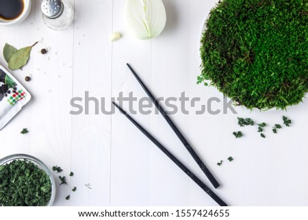 A round plastic plate with green moss stands on a white wooden table and next to it is scattered dried parsley and black chopsticks.