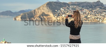 Rear view woman holding smartphone take photography of mountains hillside Calpe townscape and Mediterranean Sea. Capture moment, enjoy view tourist travelling visiting new places concept. Calpe, Spain