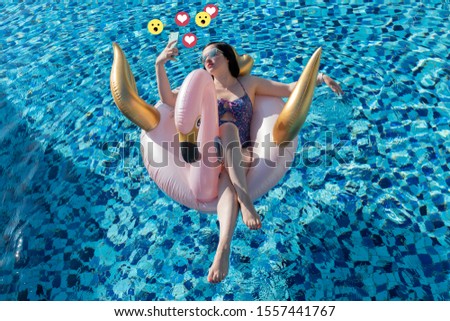 Social media influencer concept: young beautiful woman taking a selfie with mint smartphone on a swan / flamingo pool float in the blue swimming pool with heart and wow emoticons