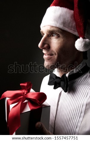 Profile portrait of young man in formal black tie tuxedo and Santa hat holding a gift with a serious expression