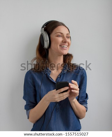 Isolated young smile face woman in blue casual jeans shirt dress in big professional headphones on head and ears listening audio podcast fm mix music from phone holding hands on white wall background Royalty-Free Stock Photo #1557515027