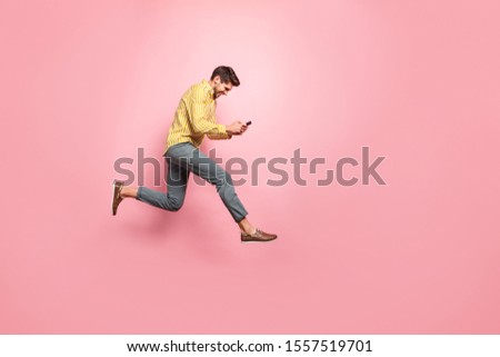 Full length profile photo of handsome guy jumping high holding telephone rushing on romantic date wear hipster striped shirt trousers isolated pink color background