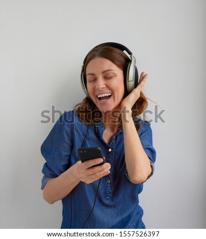 Isolated young smile face woman in blue jeans shirt dress in big professional headphones on head fix in dance on ears listening audio podcast fm mix music from phone holds in hand on white background Royalty-Free Stock Photo #1557526397