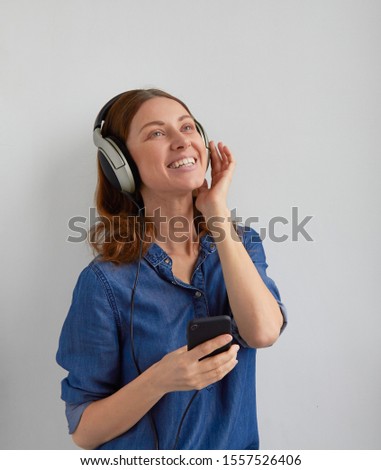 Isolated young smile face woman in blue jeans shirt dress in big professional headphones on head fix in dance on ears listening audio podcast fm mix music from phone holds in hand on white background
