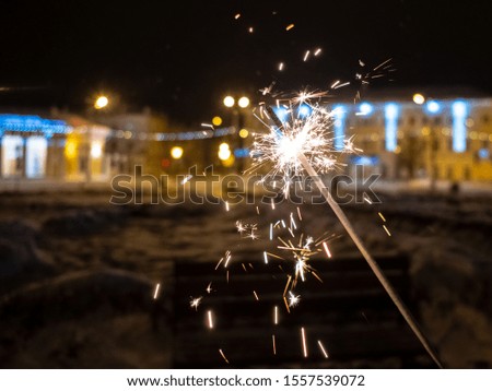 Sparkling sparkler on a city street in the dark in winter. In the background is urban festive illumination. The traditional symbol of the Christmas and New Year holidays.