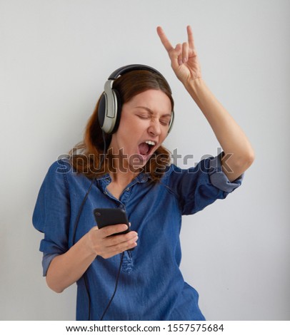 Isolated young woman in blue jeans shirt dress in big professional headphones on head dancing singing listening music show rock gesture by fingers on raised hand and holds phone on white background  Royalty-Free Stock Photo #1557575684