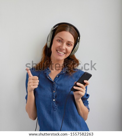 Isolated young woman with happy face and smile in blue casual jeans shirt dress in big professional headphones on head shows thumbs up and holds phone in hand on white background Royalty-Free Stock Photo #1557582263