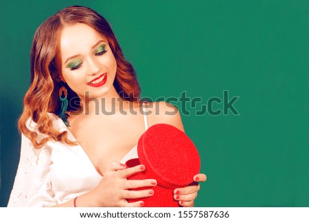 Girl with a Christmas present on a green background, opens a box. Place for text, girl with green make-up and long green earrings. White dress and long hair.
