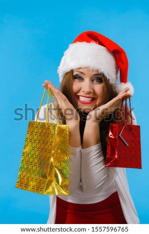 Young brunette woman is happy to give Christmas gifts. Female wearing santa claus hat holding presents festive gift bags, on blue