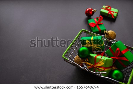 Metal shopping basket with gifts in green paper with red and yellow bows, Christmas balls of red, green and gold on a dark background. One gift and a golden ball lie side by side. Black Friday sale