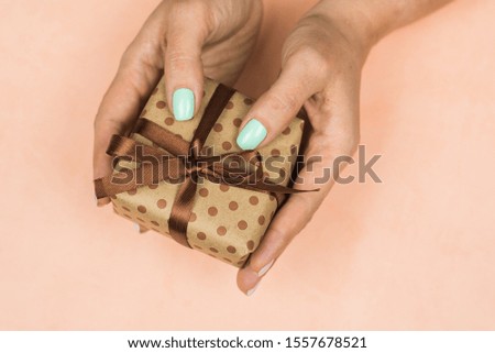 Hands of a girl with a blue pastel manicure holding a craft gift with a bow on a pink background. Concept of holiday, gifts, surprises, birthday, new year and Christmas.