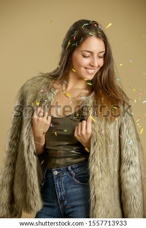 Beautiful elegant woman in fur coat under colorful confetti, party season concept, studio shot