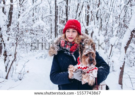 Closeup portrait of a girl with a dog in the winter forest. A woman is playing with a pet in the snow. Winter walk with a dog in the forest. Dog in a red overalls. Girl in a red knitted hat.