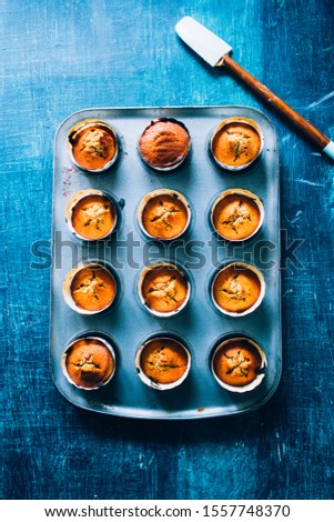 Freshly baked pumpkin's muffin lined on an aluminium baking tray  with a wooden spatula for halloween are displayed on a blu rustic board, flat lay top view,natural backlight.