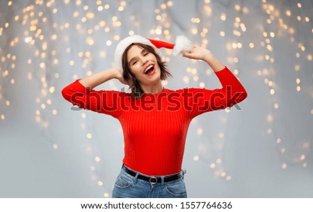 christmas, holidays and celebration concept - happy smiling young woman in santa helper hat over festive lights on grey background