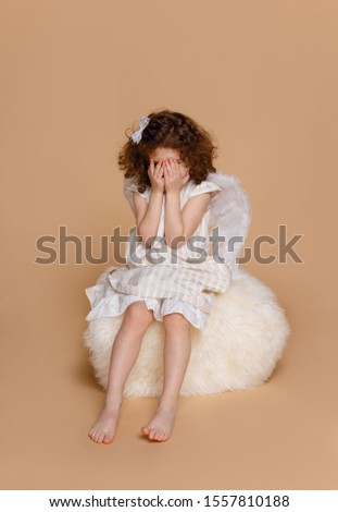 Vertical view of a little girl in wings, in dress, in studio, is hiding, over beige background.Valentine's day. Cupidon.