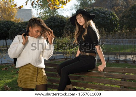 Portrait of two beautiful mexican generation z girls. A girl in a black hat sits on the back of a bench, next to a girl with curly hair and a white sweater, holds a jacket over her shoulder