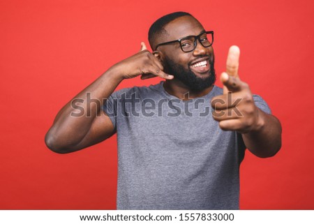 Young african american man wearing casual t-shirt standing over isolated red background smiling doing talking on the telephone gesture and pointing to you. Call me.