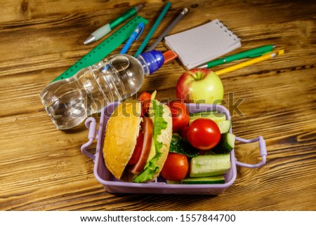 Back to school concept. School supplies, bottle of water, apple and lunch box with burgers and fresh vegetables on a wooden table