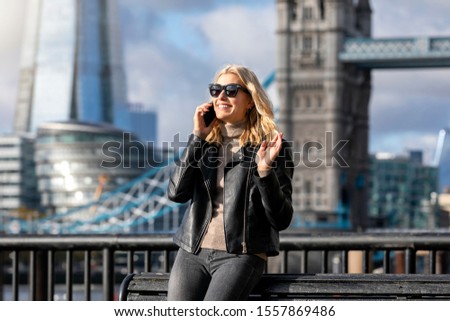 A blonde, modern city woman with sunglasses stands in front of the skyline of London, UK, and talks on her mobile phone