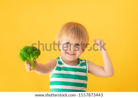 Happy child showing muscles. Portrait of strong kid with broccoli against yellow background. Healthy lifestyle concept