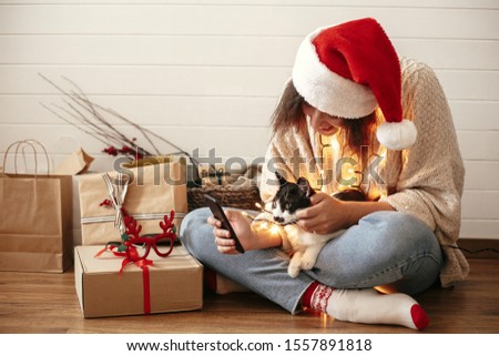 Stylish happy girl in santa hat with cute cat looking at phone screen on background of festive christmas lights and  presents. Young hipster woman hugging kitty with funny emotions