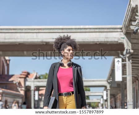 Attractive woman and afro hair with white headphones around her neck, walks down a pedestrian street, with a laptop in her right hand, on a clear sunny day