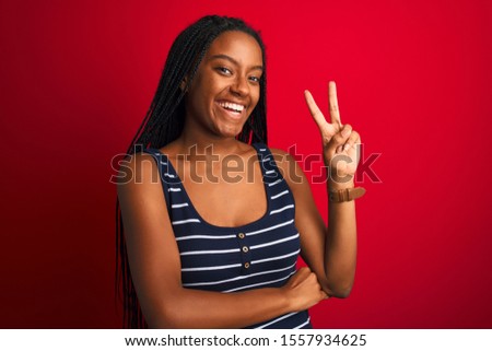 Young african american woman wearing striped t-shirt standing over isolated red background smiling with happy face winking at the camera doing victory sign. Number two.