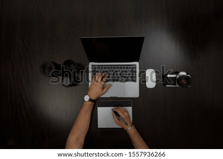Flat lay of a person editing some pictures on his laptop with a graphic design tablet  surrounded by a 35 film analog camera, and lenses. 